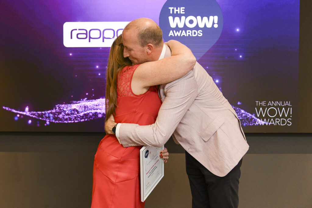 Woman in red dress holding certificate, hugging man in light coloured blazer, in front of WOW! Awards and Rapport logo.
