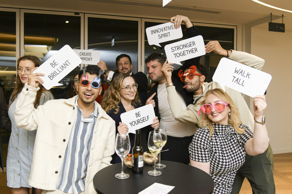Group of people wearing novelty glasses, holding up signs that read stronger together, innovative spirit, walk tall, be yourself, create rapport, and be brilliant.